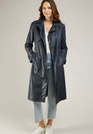 Manteau trench en cuir bleu marine avec un design croisé, de larges revers et des poches latérales ; associé à un jean clair et des baskets blanches.