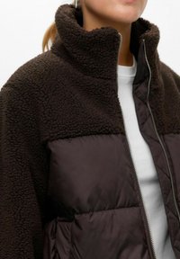 Person trägt eine dunkelbraune Fleece- und Steppjacke mit silbernem Reißverschluss über einem weißen Hemd, sichtbar von den Schultern bis zur Brust.