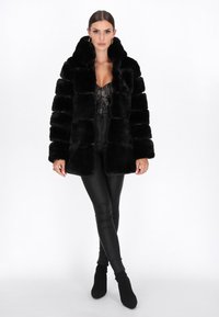 Cappotto in pelliccia sintetica nera con colletto alto, caratterizzato da strisce orizzontali e una consistenza morbida. Indossato sopra un top in pizzo nero e pantaloni neri attillati.