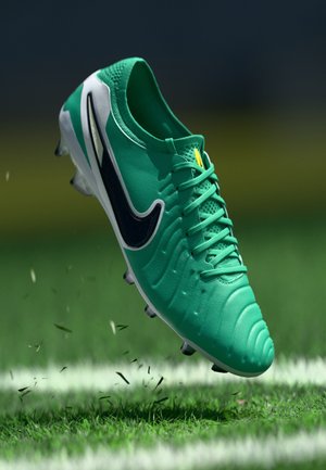 Green synthetic football boot with textured upper, black accents, white sole, and cleats. Captured mid-air over grass.