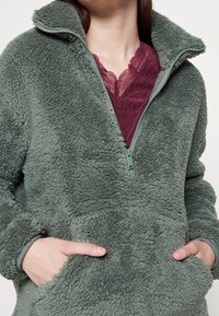 Polarowy sweter w stonowanej zieleni, z wysokim kołnierzem i pół zamkiem. Miękka, teksturowana tkanina z bocznymi kieszeniami. Nosi się go na koronkowy top.