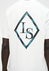 Άτομο που φοράει λευκό μπλουζάκι με μεγάλο λογότυπο σε σχήμα διαμαντιού στην πλάτη, που έχει μπλε και μαύρο περίγραμμα και γράμματα «LS» στο εσωτερικό.