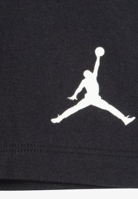 Čierne bavlnené tričko s bielym siluetovým logom skáčúcej postavy s basketbalom, umiestnené na hladkom povrchu látky.