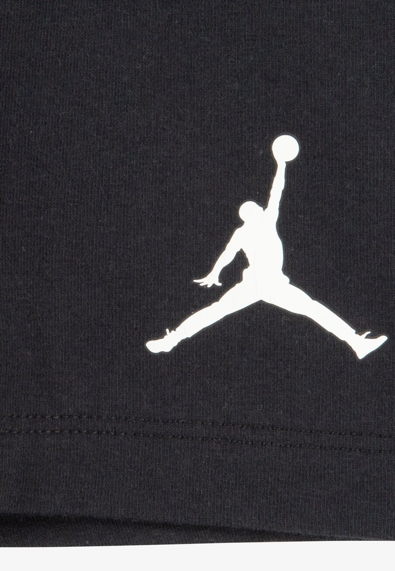 Czarny bawełniany t-shirta z białym sylwetkowym logo skaczącej postaci z koszykówką, na tle gładkiej tekstury materiału.