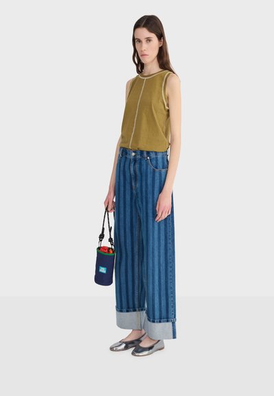 Žena stojaca v olivovom bezrukávovom topu a širokých modrých džínsach s pruhmi, drží malú tmavomodrú tašku s čiernym popruhom a červeným obsahom.