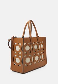 Sac fourre-tout en cuir marron avec des motifs circulaires découpés, sections en tissu couleur crème, deux poignées robustes et une forme structurée.