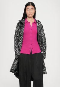 Roze gebreide cardigan met knopen, gelaagd onder een zwart-witte regenjas met zigzagpatroon. Loszittende zwarte broek maakt de look compleet.
