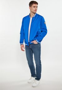 Modrá ľahká bomber bunda s bielymi pruhmi na manžetách a golieri, so zipsom vpredu, skombinovaná s priliehavými modrými džínami a bielymi teniskami.