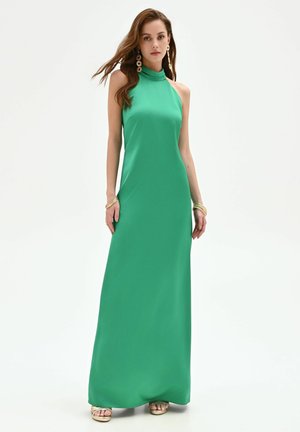 Femme portant une robe verte longue sans manches, des boucles d'oreilles en or, des bracelets et des sandales à plateforme, posant sur un fond blanc.