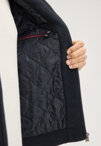 Donkerblauwe jas met een gevoerd zwart binnenwerk. Bevat een ritszak met rode rand en een geribbelde zoom. Hand zichtbaar die de jas vasthoudt.