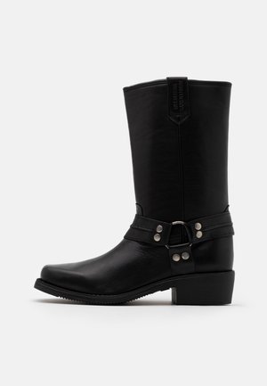 Stivale da motociclista in pelle nera fino a metà polpaccio con cinturino a imbracatura borchiato argento e tacco basso a blocco su sfondo chiaro uniforme.