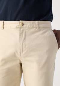 Beige katoenen chino's met een knoopsluiting, een vlakke voorkant en zijzakken. Gladde textuur en een getailleerde pasvorm, gedeeltelijk bedekt door een donker shirt.