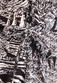 Tissu à motifs noir et blanc avec un détail nœud papillon, fermeture à boutons et design texturé, présentant des formes et des lignes abstraites.