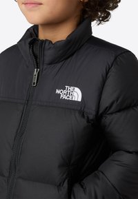 Svart isolert jakke med høy krage, frontglidelås og hvit logo. Stoffet ser glatt ut med en puffet tekstur.