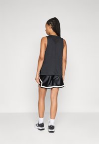 Kvinna med flätat hår, iklädd svart ärmlös topp, svarta sportshorts med vit trim, vita strumpor och svart-vita sneakers, står upp.