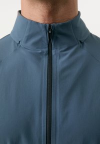 Herenjack in een gedempte blauwe stof; heeft een hoge kraag en een centrale ritssluiting met contrasterende zwarte rits. Soepele textuur.