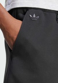 Černé sportovní kalhoty s hladkým povrchem, s boční kapsou a vyšitým trojlístkovým logem ve tmavě šedé barvě na přední straně.