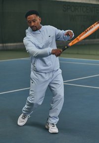 Gaiši zils sporta tērps ar jaku ar rāvējslēdzēju un biksēm ar elastīgu jostasvietu, valkāts kopā ar baltiem sporta apaviem, turēts tenisa rakets.