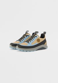 Lys blå og beige sneakers med svarte detaljer, teksturerte paneler og tykke såler, med svarte snørebånd og en krok bak hælen.