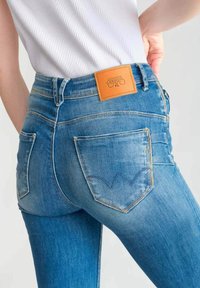 Jeans en denim bleu avec une taille haute, présentant une patch en cuir beige au dos de la ceinture et deux poches arrière avec des accents de couture.