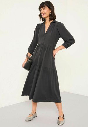 Schwarz, langärmliges Kleid mit V-Ausschnitt, tailliertem Oberteil und gestuftem Rock. Kombiniert mit grauen, flachen Schuhen und einer kleinen schwarzen Tasche.