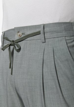 Lysegrå bukser med læg, knaplukning, bæltestropper og et bundet stofsnøre i taljen båret med en hvid skjorte.