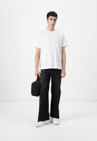 Witte t-shirt met een klein, multicolored logo, gecombineerd met zwarte flared jeans en witte sneakers. Houdt een kleine zwarte tas vast.