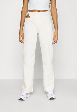 Pantalons larges de couleur crème en coton avec une ceinture asymétrique et un détail de ceinture. Portés avec des baskets.