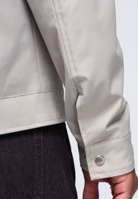 Manche et poignet d'une veste beige clair avec détail de bouton-pression, portée sur un pantalon en tissu foncé texturé.