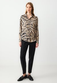 Beige blouse met hoge kraag en knopen, versierd met zwarte zebra-strepen, gecombineerd met zwarte tapered broek en zwarte instappers. Smooth, lichtgewicht stof.
