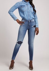 Camisa de mezclilla en azul claro con botones al frente y dos bolsillos en el pecho, combinada con jeans ajustados desgastados con rodillas rasgadas y parches en azul oscuro.