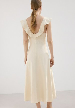 Kobieta o długich brązowych włosach związanych nisko w kucyk, ubrana w kremową, bezrękawnikową sukienkę z falbanką i głębokim dekoltem na plecach, stoi na tle jednolitego, jasnego tła.