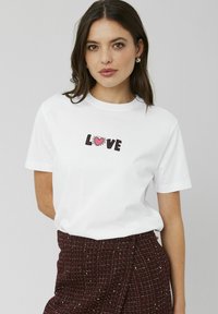 T-shirt en coton blanc avec "LOVE" en lettres noires en gras et un cœur rose, manches courtes et col rond.