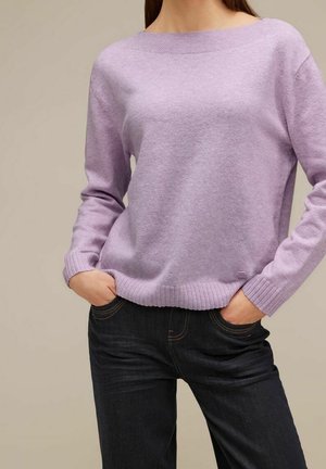 Femme portant un pull lilas à manches longues avec poignets et ourlet côtelés, mains dans les poches d’un jean bleu foncé, sur un fond uni.