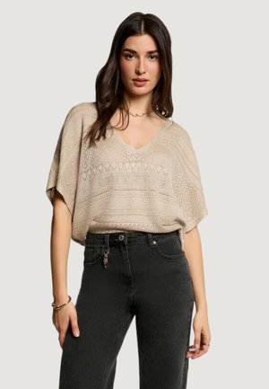 Kobieta ubrana w beżowy, wzorzysty sweter z dekoltem w serek oraz wysokogabarytowe czarne dżinsy, stojąca na tle jednolitego, jasnego tła.