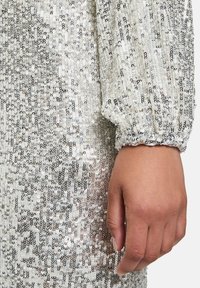 Srebrny materiał sukienki pokryty cekinami z odbijającymi się cekinami, balonowe rękawy i marszczone mankiety, na zdjęciu widoczna ręka trzymająca materiał.