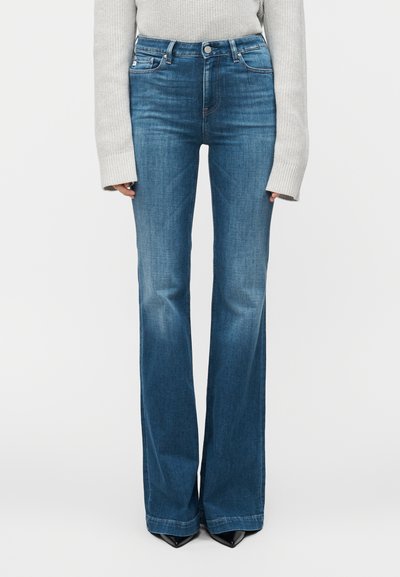 Μπλε καμπανέ τζιν από denim, με ψηλή μέση, σχεδιασμό με πέντε τσέπες και διακριτική ξεθώριαση. Φορεμένο με ανοιχτό γκρι πλεκτό πουλόβερ.