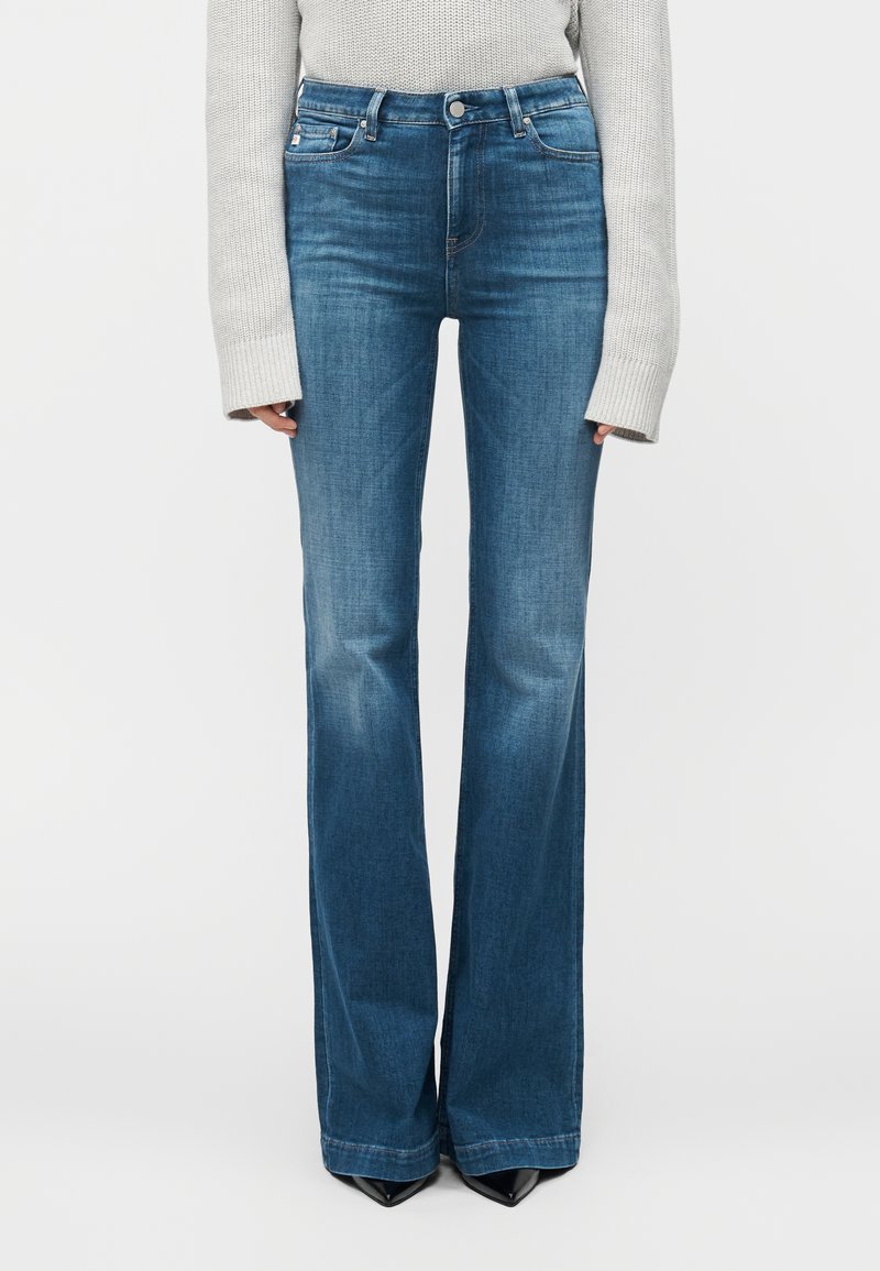 Blå utsvängda jeans i denim med hög midja, fem fickor och subtila blekningsdetaljer. Bärs med en ljusgrå stickad tröja.