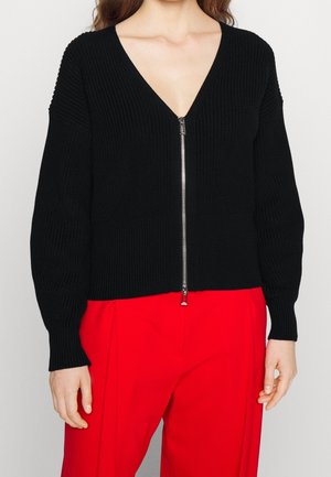 Femme portant un pull en maille noir zippé avec un col en V et un pantalon rouge taille haute, debout devant un fond uni.