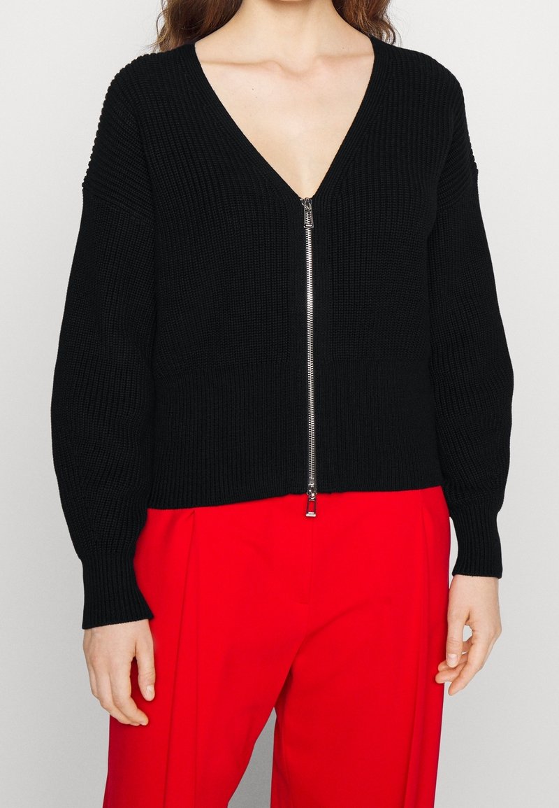 Femme portant un pull en maille noir zippé avec un col en V et un pantalon rouge taille haute, debout devant un fond uni.