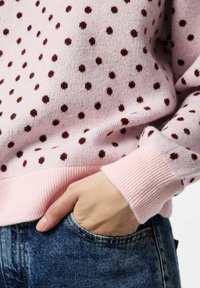 Pull en tricot rose clair avec des pois marron foncé. Poignets et ourlet côtelés, texture douce, coupe décontractée, avec une main partiellement dans une poche en denim bleu.