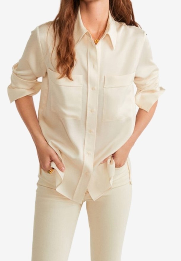 Femme portant une chemise crème boutonnée avec manches retroussées et pantalon assorti, mains dans les poches de manière décontractée, sur fond blanc.