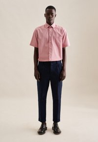 Korte mouw, lichtroze overhemd met knoopjes en een borstzak, gecombineerd met marineblauwe broeken en zwarte schoenen, staande op een neutrale achtergrond.