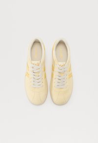 Gelbe Sneaker mit cremefarbenen Akzenten, runder Zehenkappe und weißen Schnürsenkeln. Besitzt eine glatte Oberfläche und ein dezentes Logodetail auf der Zunge.