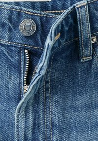 Gros plan sur un jean en denim bleu montrant une fermeture éclair en métal ouverte, un bouton en métal avec du texte, et une boucle de ceinture avec une couture beige.