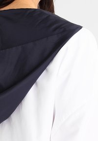 Kurtka z kapturem z granatowym kapturem i białym korpusem. Materiał wydaje się gładki i lekki, z widocznymi detalami przeszyć.