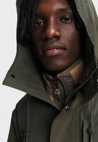 Gros plan d'une personne portant une veste à capuche vert olive sur une polaire beige zippée avec des cordons visibles.