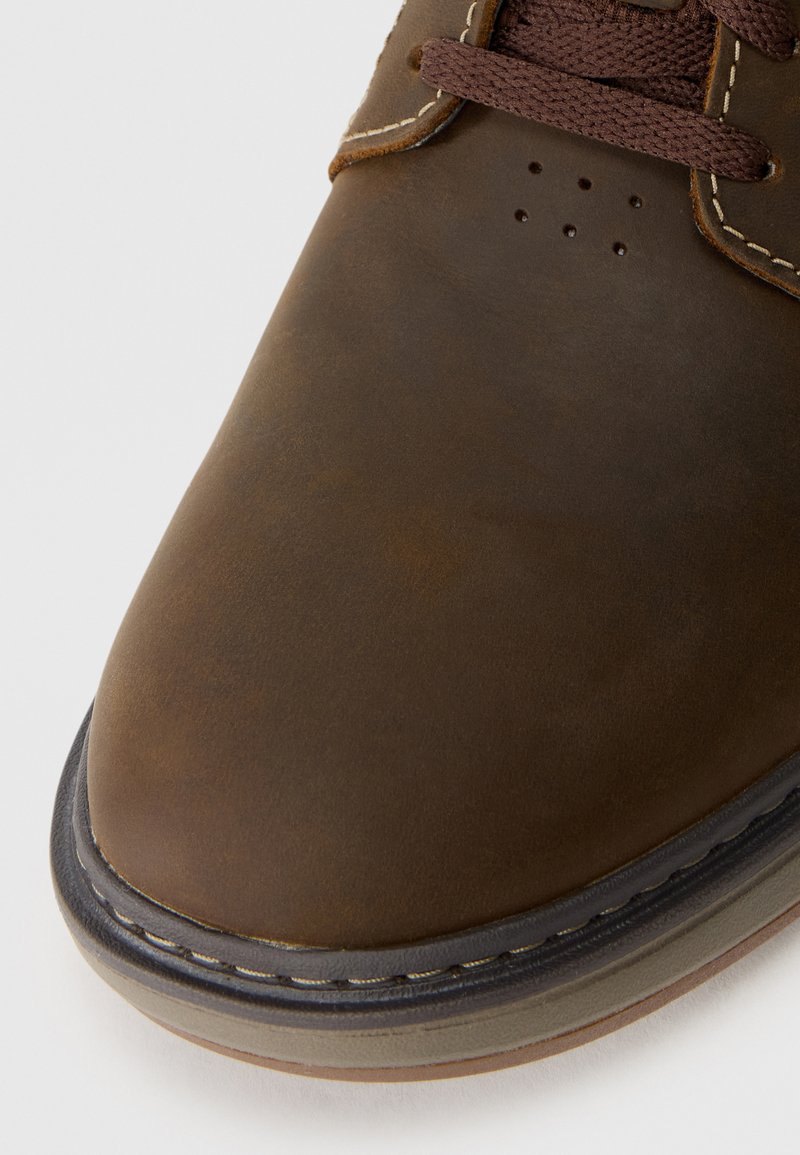 Zapato de cuero marrón con una superficie lisa, punta redondeada, cordones marrones y pespuntes en contraste. Presenta orificios de ventilación cerca de los cordones.