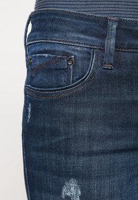 Tmavě modré džíny z denimu se zadní kapsou, viditelným šitím, mírným opotřebením a hladkým povrchem.