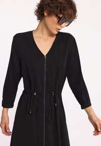 Czarna sukienka z dekoltem w kształcie litery V, długimi rękawami i zamkiem z przodu. Zawiera regulowany sznurek w talii oraz gładką teksturę materiału.